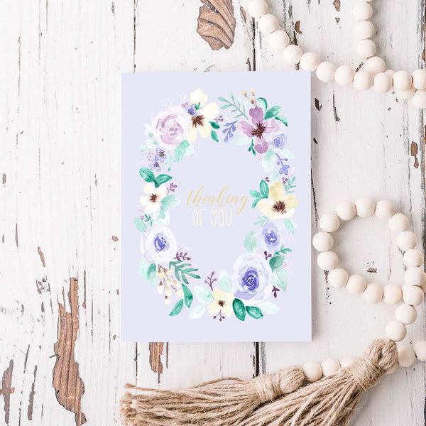 watercolor wild flower wreath on a friendship greeting card that says thinking of you in the center with a white A2 envelope shown laying on a rustic white wood table with a white wood bead garland