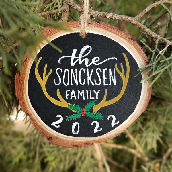 Hand painted rustic wood slice ornament personalized with family name and the year with painted deer antlers and evergreen branches and red berries