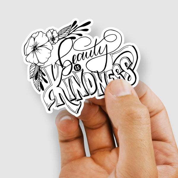 Hand lettered illustrated 3" vinyl sticker that says beauty is kindness with floral illustration in black and white shown with a woman's hand holding the sticker