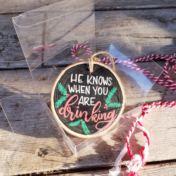 Wood slice ornament that says he knows when you are drinking with evergreen and berry illustrations in red green and white in a clear gift box with red and white string