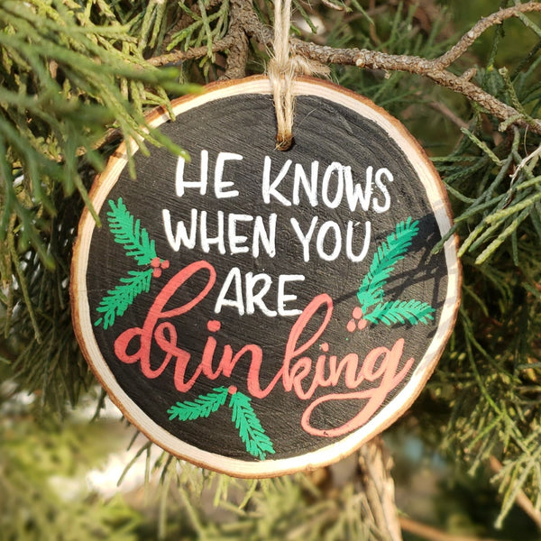 Wood slice ornament that says he knows when you are drinking with evergreen and berry illustrations in red green and white hanging in a tree