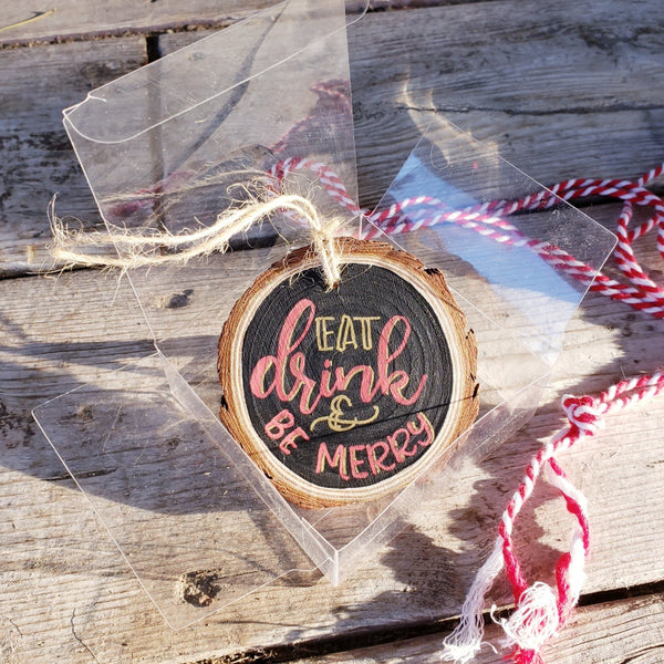 rustic wood slice ornament that says eat drink & be merry in gold and red hand lettering in a clear gift box with red and white string