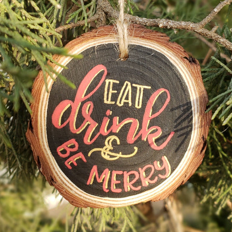rustic wood slice ornament that says eat drink & be merry in gold and red hand lettering hanging in a tree