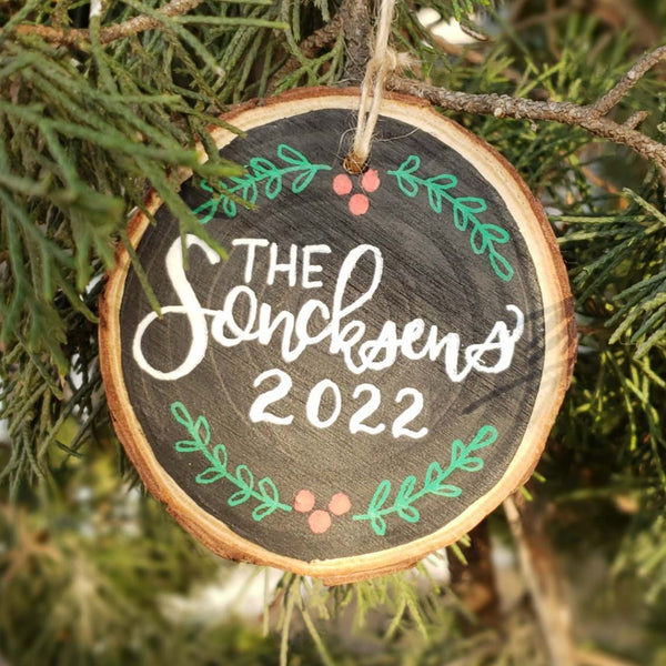 Hand painted rustic wood slice ornament personalized with family name and year in hand lettering and red and green foliage doodles