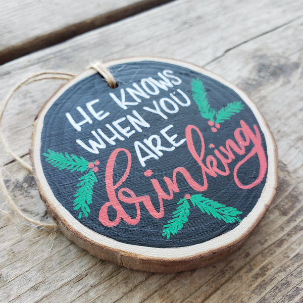 Wood slice ornament that says he knows when you are drinking with evergreen and berry illustrations in red green and white