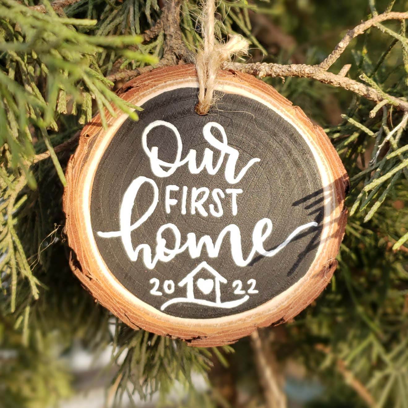 Hand painted rustic wood slice ornament that says our first home with the year in hand lettering and a little house doodle