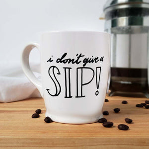 16 oz hand painted white ceramic coffee mug that says i don't give a sip in black hand lettering shown with scattered coffee beans and a french press and floursack towel in the background