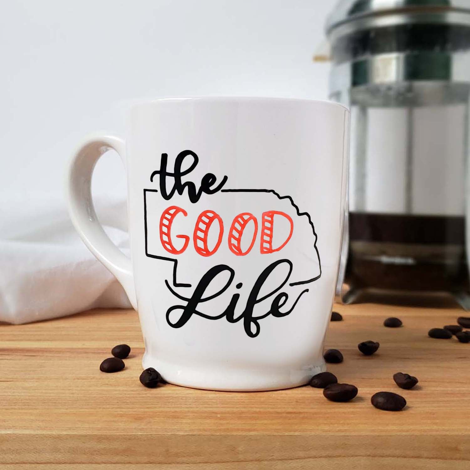 16 oz hand painted white ceramic coffee mug that says the good life with the outline of the state of Nebraska in red and black hand lettering shown with scattered coffee beans and a french press and floursack towel in the background