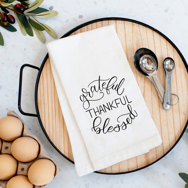 White floursack towel with black hand illustrated design that says grateful thankful blessed shown folded on a serving tray with a set of measuring spoons and fresh eggs