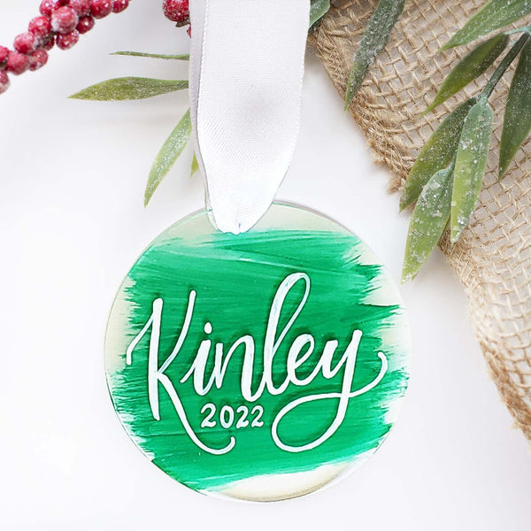 Round clear acrylic ornament personalized with first name and year in hand lettering with a white silk ribbon and green paint strokes in the background