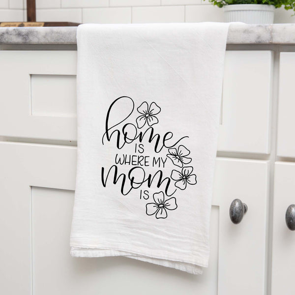 White floursack towel with black hand lettered illustrated design that says home is where my mom is with flower doodles shown folded and hanging from a countertop in a modern kitchen