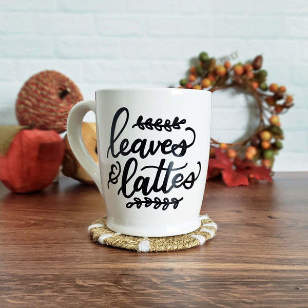 Hand painted white ceramic mug that says leaves & lattes with sprigs of leaves doodles shown sitting on a wooden table with fall autumn decor in the background