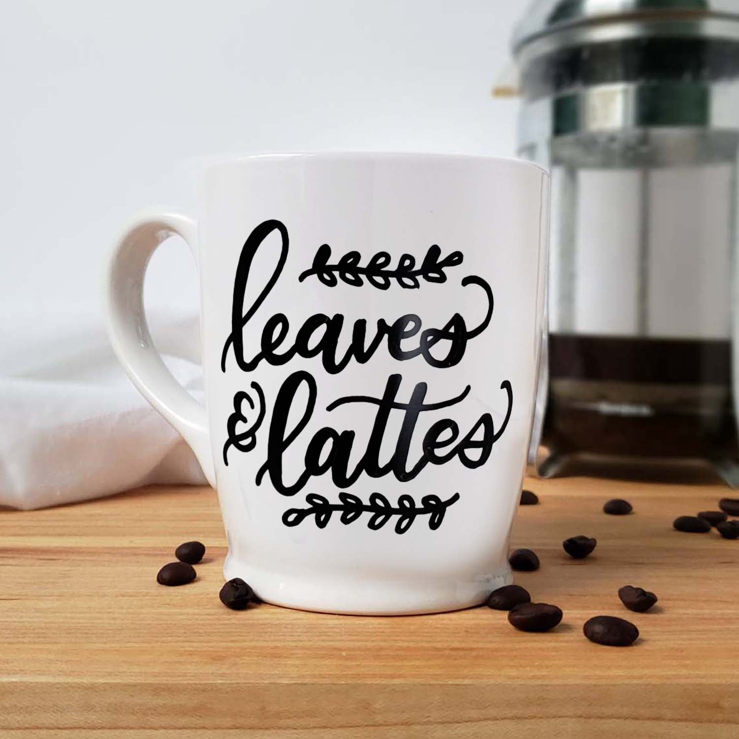 Hand painted white ceramic mug that says leaves & lattes with sprigs of leaves doodles shown on a kitchen counter with a french press, towel and scattered coffee beans