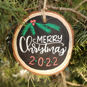 Hand painted rustic wood slice ornament that says Merry Christmas 2022 in hand lettering and evergreen and berry doodles