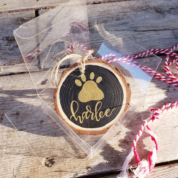 rustic wood slice ornament that says your pet's name hand lettered in gold with a paw print in gold in a clear gift box