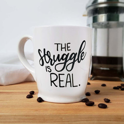 16 oz hand painted white ceramic coffee mug that says the struggle is real in black hand lettering shown with scattered coffee beans and a french press and floursack towel in the background
