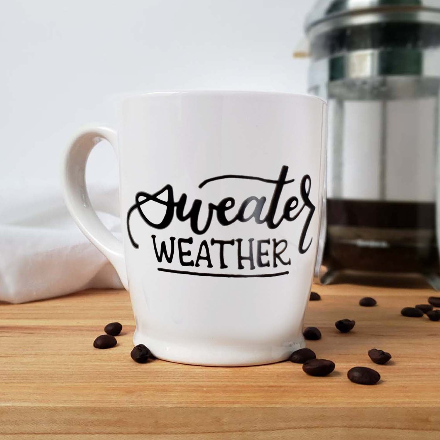 Hand painted white ceramic mug that says sweater weather in black hand lettering shown on a kitchen counter with a french press, towel and scattered coffee beans